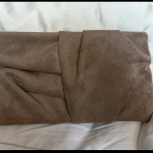 Brun mocka kuvertväska med kedja - Snygg brun kuvertväska i mocka med stilren omlott-design. Väskan har en silverfärgad kedja som axelrem och ett magnetlås. Insidan har ett fack med dragkedja för småsaker. Perfekt storlek för det viktigaste och en clean look.