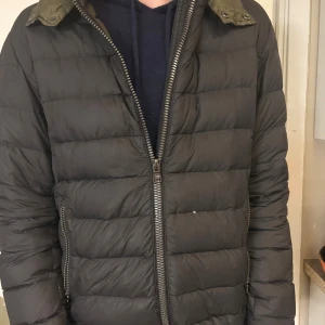 Svart Colmar pufferjacka med huva - Svart pufferjacka från Colmar med huva och grov dragkedja framtill. Jackan har ett quiltat mönster och en rund Colmar-logga på ärmen. Fickor med dragkedja på sidorna och långärmad modell. Perfekt för kalla dagar och riktigt snygg streetstil. Lagad på vänster armbåge (se bild 4) har även litet hål vänster om dragkedjan framtill (se bild 5)