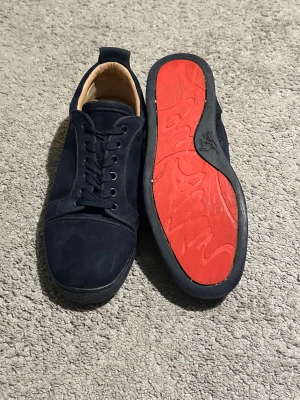 Mörkblå Christian Louboutin skor - Säljer ett par mörkblå sneakers från Christian Louboutin i mocka med klassisk röd sula och svarta snören. Skorna har rund tå, platt sula och beige läderfoder. Perfekta för dig som vill ha en stilren och lyxig look med en ikonisk touch.