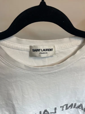 Vit Saint Laurent t-shirt med tryck - Vit t-shirt från Saint Laurent med klassiskt svart logotryck på bröstet. Modellen har rund hals och korta ärmar, tillverkad i mjuk bomull. Perfekt för dig som gillar stilrena designer och vill ha ett statement-plagg i garderoben. Passar som XS
