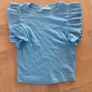 Blå ribbad topp med volangärmar från Zara - Säljer en ljusblå topp från Zara i ribbad bomull med korta volangärmar. Modellen är normal i passningen och har rund hals. Perfekt för dig som gillar en söt och trendig look med extra detaljer på ärmarna.
