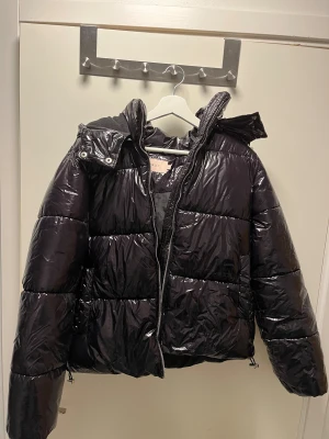Svart glansig pufferjacka från Nelly - Svart pufferjacka från Nelly med glansig finish, huva och dragkedja framtill. Jackan är vadderad och har en oversized look med långa ärmar. Perfekt för kalla dagar när du vill ha både stil och värme. Andvänt få tal gånger max 3gånger, ny pris 800kr. Hör av er om ni har mer frågor!🤗