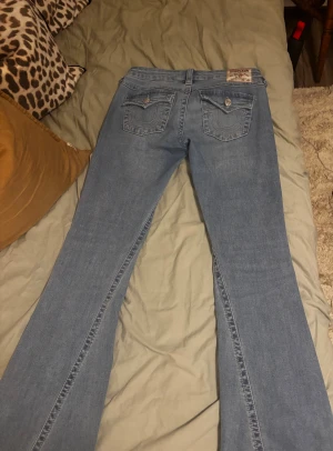 True Religion Joey ljusblå flare jeans - Säljer ett par ljusblå True Religion Joey jeans med låg midja och flare-ben. Klassiska jeansfickor med dekorativa sömmar och silverfärgade knappar bak. Mjukt denim-material som sitter snyggt och har en cool vintage-vibe.