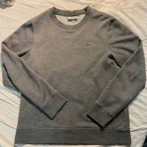 Grå sweatshirt från Lacoste - Snygg grå sweatshirt från Lacoste med klassisk krokodillogo broderad på bröstet. Tröjan har rund hals, långa ärmar och ribbade muddar vid ärmslut och nederkant. Perfekt för chill dagar och enkel att matcha med jeans eller joggers.