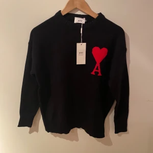 Svart stickad tröja från Ami Paris - Svart stickad tröja från Ami Paris med röd hjärtapplikation och bokstaven A på bröstet. Tröjan har rund hals, ribbade muddar och lång ärm. Perfekt för dig som gillar stilrena plagg med en twist. Snygg att matcha med jeans eller kjol.