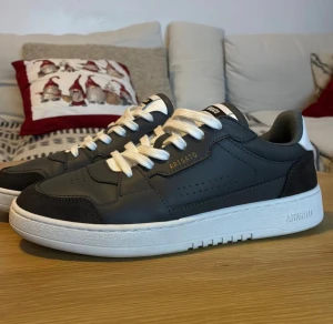 Axel Arigato Area Lo - Ett par Axel Arigato Area Lo sneakers i nyskick. Skorna har en vit sula och grå ovandel med detaljer i mörkgrå mocka.  Skorna är i nyskick. Nypris 2800:-