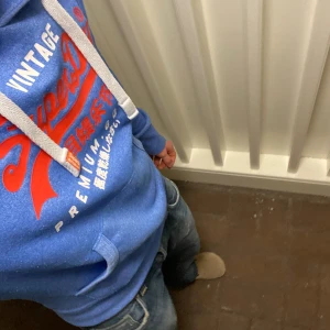 Superdry Hoodie - Snygg blå hoodie från Superdry med stort rött och vitt tryck på bröstet och japanska tecken. Tröjan har huva med vita snören, känguruficka och ribbade muddar. Perfekt för dig som gillar streetwear och vill ha en cool look. Den är en S men den sitter som en M. Om du undrar något är det bara att fråga🙌✅