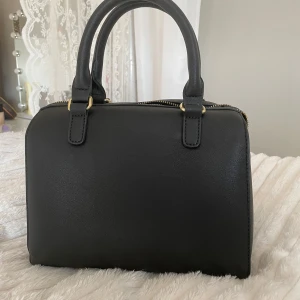 Svart handväska från Don Donna - Stilren svart handväska från Don Donna med rundad form och två rejäla handtag. Väskan har guldfärgade metalldetaljer och en snygg dragkedja upptill. Materialet är slätt syntet som ger en clean look. Perfekt storlek för det viktigaste och passar till många outfits.