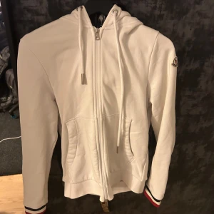 Vit hoodie från Moncler med dragkedja - Säljer en vit hoodie från Moncler med hel dragkedja, huva med snörning och fickor framtill. Tröjan har Moncler-logga på ärmen och coola rödblå ränder på muddar och insidan av huvan. Perfekt för dig som gillar stilrena och sportiga plagg.