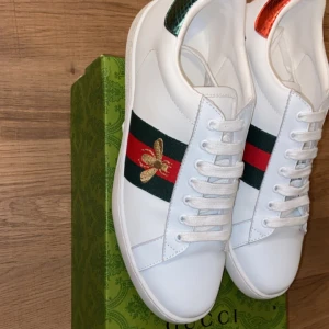 Gucci Ace vita sneakers med broderad bi - Gucci Ace sneakers i vitt skinn med klassiska gröna och röda ränder på sidan, broderad guldfärgad bi och metallic-detaljer i rött och grönt på hälen. Rund tå, snörning och platt sula med unikt mönster undertill. Snygga och lyxiga sneakers för dig som gillar statement.