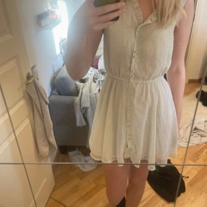 Vit prickig kort klänning med knappar - Supersöt vit kort klänning med svarta prickar, knappar hela vägen framtill och markerad midja. Ärmlös modell i lätt och luftig polyester, perfekt för varma dagar. Klänningen har en klassisk krage och ett flowy fall som ger en chill vibe.