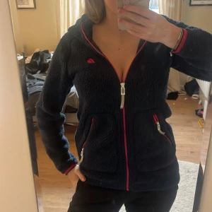 Mörkblå fleecejacka från Didriksons - Mysig mörkblå fleecejacka från Didriksons med huva och rosa detaljer. Jackan har hel dragkedja framtill, två fickor med röda dragkedjor och broderad logga på bröstet. Perfekt för kyliga dagar och enkel att slänga på sig över en hoodie.