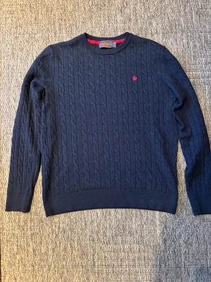 Marinblå kabelstickad tröja Morris - Snygg marinblå kabelstickad tröja från Morris i 100% merinoull. Rund hals, långärmad och med röd broderad logga på bröstet. Perfekt för dig som gillar klassisk stil med en modern twist. Storlek m men passar s