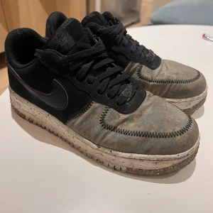 Nike Air Force 1 svart/grå sneakers - Nike Air Force 1 sneakers i svart och grått med färgstänk på textildelen och en robust, prickig sula. Skorna har snörning, rund tå och klassisk Nike-logga på sidan och hälen. Materialmix av skinn och textil, med tydliga kontrastsömmar runt tån.