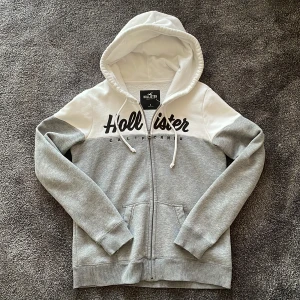 Grå och vit hoodie från Hollister - Snygg hoodie från Hollister i storlek S, med grå och vit färgblock-design. Tröjan har huva med snörning, dragkedja framtill och broderad Hollister-logga över bröstet. Tillverkad i mjuk bomullsmix, perfekt för en avslappnad stil.