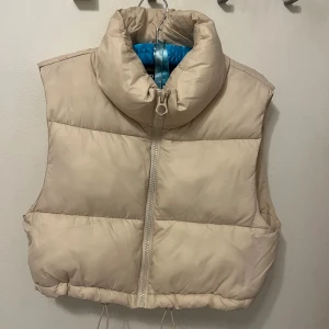 Beige dunväst från Stradivarius - Säljer en beige dunväst från Stradivarius med hög krage och grov dragkedja framtill. Västen har quiltad design och dragsko nedtill för att justera passformen. Perfekt att slänga över en hoodie för extra värme under kyliga dagar. Västen har en liten effekt knappt synlig på vänster sida.