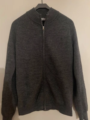 Mörkgrå kofta med dragkedja från Sandö - Snygg mörkgrå kofta från Sandö i storlek M. Koftan är stickad och har en hel dragkedja framtill samt ribbad krage och muddar. Perfekt för lager-på-lager och chill dagar. Enkel design som funkar till många stilar. Inga skador på den.
