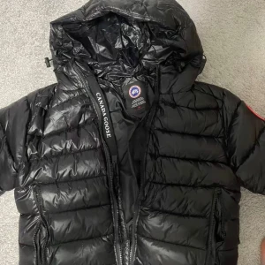 Svart pufferjacka från Canada Goose - Säljer en svart pufferjacka från Canada Goose med huva och dragkedja. Jackan har det klassiska Canada Goose-märket på ärmen och är tillverkad i glansig polyester. Perfekt för kalla dagar och har en snygg, modern look.