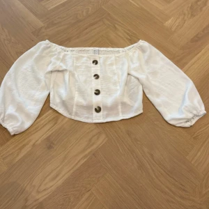 Vit offshoulder blus från Amisu - Supersöt vit blus från Amisu i storlek S. Offshoulder-modell med elastisk kant upptill och långa puffärmar. Framtill sitter fem snygga knappar i brunmelerad ton. Blusen är croppad och gjord i lätt bomullstyg, perfekt för varma dagar.