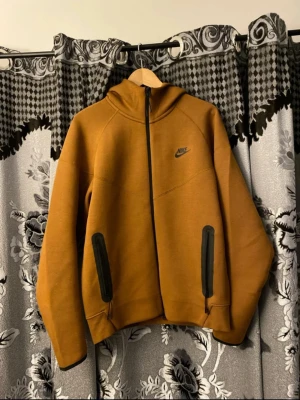 Nike Tech Fleece set i brun - Nike Tech Fleece set i snygg brun färg. Hoodien har dragkedja, huva och svarta detaljer vid fickorna. Byxorna har smal passform, svarta dragkedjor på fickorna och snörning i midjan. Byxorna är i storlek S och Hoodien är i storlek M