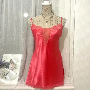 Röd-Orange satin nattlinne från Victoria’s Secret - Snygg kortklänning i röd-orange färg med spetsdetaljer vid halsringning och nederkant samt en öppen rygg med knytband. Storlek: M, passar även S-L (36-40) då du kan justera bandet på baksidan. Dockan är i storlek 36.