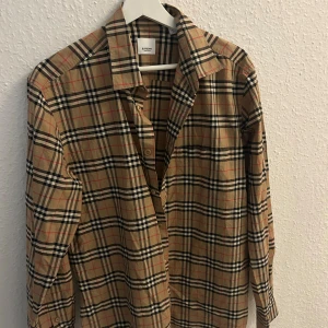 Burberry rutig skjorta i bomull - Snygg klassisk skjorta från Burberry i beige med ikoniskt rutigt mönster i svart, vitt och rött. Skjortan har lång ärm, knappar framtill och mjukt bomullsmaterial. Perfekt för dig som gillar stilrena och tidlösa plagg med lyxig känsla.