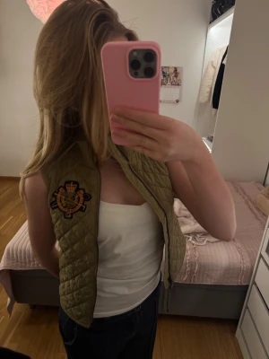 Quiltad beige/grön väst med broderad emblem - Snygg quiltad beige/mossgrön vintage  väst från Ralph Lauren med broderad emblem på bröstet. Västen har hög krage, dragkedja framtill och coolt rutmönster. En speciell kollektion som tillhör just ridning. Den har ett litet lättlagat hål vid armhålan kanten därav priset💕