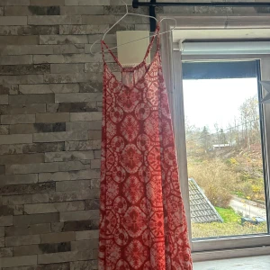 Röd mönstrad långklänning Rainbow XL - Säljer en färgstark långklänning från Rainbow i storlek XL. Klänningen har tunna axelband och ett coolt rött och vitt mönster som ger den en unik vibe. Materialet är mjukt och lätt polyester, perfekt för varma dagar. V-ringad framtill och loose passform.