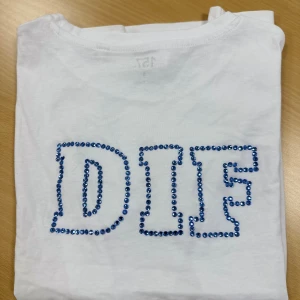Vit DIF t-shirt med blå paljetter på ryggen  - Snygg vit t-shirt med DIF-text baktill, dekorerad med blå paljetter. Rund hals och klassisk passform, perfekt för dig som vill visa ditt lagstöd med stil. Tröjan är i grund från lager 157, går att tvätta OBS! Bara i 30 grader, ingen torktumling 