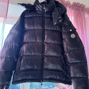 Svart Moncler pufferjacka med huva - Svart pufferjacka från Moncler med ikonisk logga på ärmen, dragkedja framtill och praktisk huva. Jackan har quiltad design och innerfoder med serietidningsmönster. Materialet är glansig polyester och dunfyllning, perfekt för kalla dagar. Ingen aning om äkthet. Kom med ett bud. 