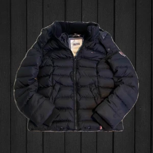 Svart jacka från Hilfiger denim  - Säljer en riktigt skön jacka från Hilfiger denim  med huva och ribbade muddar. Jackan har dragkedja framtill och snygga Hilfiger detaljer på ärmen. Perfekt för kalla dagar och har en cool och avslappnad look. Det finns en liten defekt på ärmen men syns knappt. Hör av dig vid funderingar.