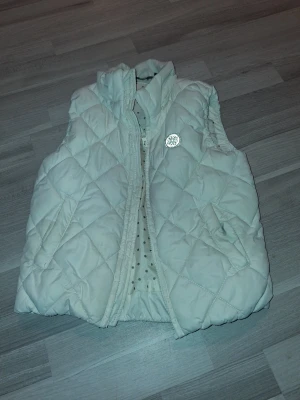 Vit quiltad dunväst från Lindex - Barn vit dunväst från Lindex, Out Wear-kollektionen. Västen har hög krage, två fickor och dragkedja framtill. Insidan är fodrad med prickigt tyg i beige och brunt. Reflekterande snöflinge-detalj på bröstet och ryggen. Perfekt för kyligare sommar-vår-höst dagar. Använd men utan synligt och påverkad slitage. Saknar luvan. Storlek 134/140