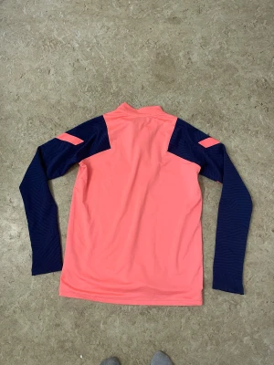 Nike Tottenham långärmad träningströja - Snygg långärmad träningströja från Nike x Tottenham Hotspur i en fräsch rosa färg med marinblå ärmar och detaljer. Tröjan har half zip framtill, klubbmärke på bröstet och är gjord i mjukt polyester-material som andas. Perfekt för dig som gillar fotboll och vill sticka ut på planen.