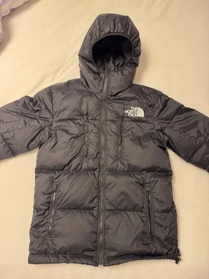 Svart dunjacka från The North Face S - Svart dunjacka från The North Face med huva och broderad logga på bröstet och ryggen. Jackan har flera dragkedjeförsedda fickor och är vadderad med 550 fill power dun. Perfekt för kalla dagar och har en clean look. Den är i storlek S. Jag har personligen knappast använt den och den är relativt ny. Köpte den 2800kr