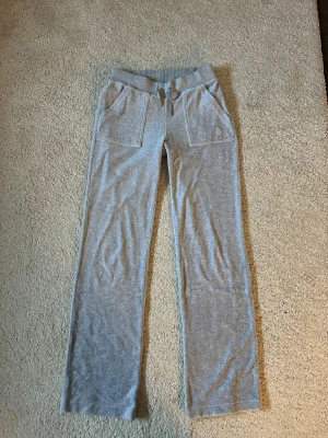 Grå mjukisbyxor Juicy Couture, XS - Grå snygga och trendiga mjukisbyxor från Juicy Couture i storlek XS. Byxorna har bred resår i midjan med snörning, stora framfickor och en bakficka med lock och tryckknappar.