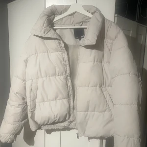 Beige pufferjacka från 157 - Säljer en beige pufferjacka från 157 med hög krage och dragkedja framtill. Jackan är quiltad med breda kanaler och har en lite croppad passform. Perfekt för kalla dagar när du vill vara både varm och snygg.