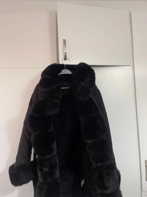 Svart parkajacka med fuskpälsdetaljer - Säljer en längre svart vinterjacka med huva med fluffig fuskpäls runt krage och ärmslut, jackan har dragkedja,  pälsen som finns vid huvan och på sidorna vid dragkedjan kan man ta bort. Är storkek S funkar som M också skulle jag säga använd en säsong säljer för att den tyvärr är för liten för mig.  Finns även snörning i midjan samt två fickor med dragkedja. 