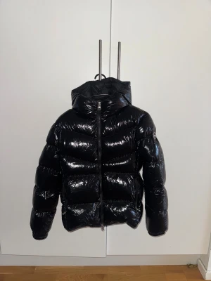 Svart glansig dunjacka från Colmar - Säljer en svart, glansig dunjacka från Colmar med huva och dragkedja framtill. Jackan har en puffad look och är tillverkad i syntetmaterial med dunfyllning. Perfekt för dig som vill ha en varm och snygg jacka med cool streetstil.