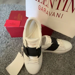 Valentino Garavani Open sneakers vit/svart - Säljer ett par snygga Valentino Garavani Open sneakers i vitt skinn med bred svart rem över mitten. Klassisk rund tå, platt sula med nitar och vita snören. Märkeslogga på plösen och cool kontrast mellan färgerna. Perfekta för dig som gillar exklusiv streetstyle.