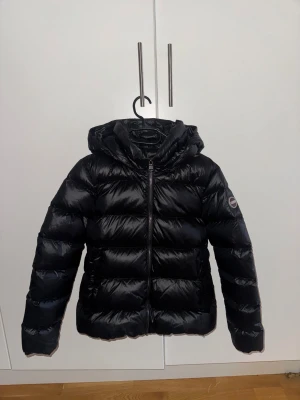 Svart dunjacka från Colmar med huva - Svart pufferjacka från Colmar med huva och dragkedja framtill. Jackan har quiltad design, två stora fickor på insidan och ett märke på ärmen. Fylld med naturligt dun för extra värme. Perfekt för kalla dagar och har en clean, modern look. Avtagbar luva