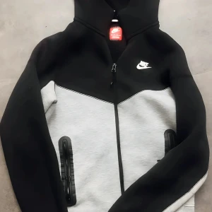 Nike hoodie svart och grå med dragkedja - Nike hoodie i svart och ljusgrå färg med hel dragkedja framtill och huva. Snyggt tvåfärgad design med vita Nike-loggan på bröstet. Praktiska fickor med svarta detaljer och långärmad modell. Perfekt för chill eller träning. Priset 699