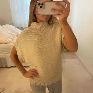 Beige stickad polotröja  - Säljer en beige stickad polotröja med hög krage och bred passform. Tröjan är ärmlös och har en chunky stickad struktur som ger en mysig och trendig look. 