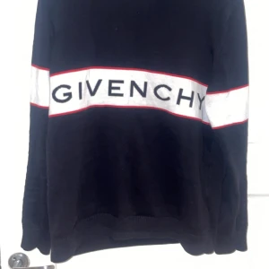 Svart stickad tröja Givenchy - Svart stickad tröja från Givenchy med vit och röd rand över bröstet och ryggen. Stor logotyp framtill. Tröjan har långärmad passform och ribbade muddar vid ärmslut och nederkant. Perfekt för dig som gillar streetstyle och designer.