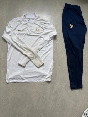 Nike France tracksuit - Snygga marinblå träningsbyxor från Nike med Frankrikes landslagsemblem i guld på höften. Byxorna har elastisk midja, smal passform och diskret mönster på nedre delen av benen. Perfekta för fotboll eller chill. Materialet är polyester.