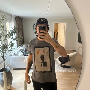 Grå Basquiat x Neuw t-shirt med print - Säljer en grå t-shirt från Jean-Michel Basquiat x Neuw med ett stort konstnärligt print på framsidan. Trycket har motiv i svart, rött och gult med texten '1960'. T-shirten har rund hals och korta ärmar, tillverkad i mjuk bomull. Perfekt för dig som gillar street och konst.