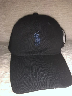 Svart Polo Ralph Lauren keps - Svart keps från Polo Ralph Lauren med klassisk böjd skärm och blå broderad logga framtill. Justerbar rem med metallspänne baktill för perfekt passform. Snygg och stilren design som passar till allt. Materialet är bomull.