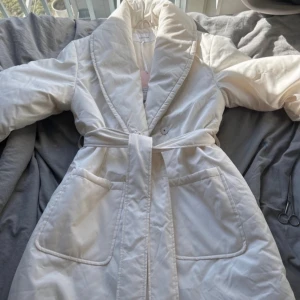 Vit quiltad pufferjacka med bälte - Säljer en vit pufferjacka med quiltad design och bred krage. Jackan har stora fickor framtill och ett matchande bälte i midjan för snygg passform. Perfekt för kyliga dagar och har en modern, clean look som är lätt att styla med olika outfits.