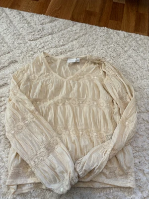 Beige blus med spets från Vila - Säljer en romantisk beige blus från Vila i storlek XS. Blusen har v-ringning, långa ärmar och är dekorerad med breda spetsband. Materialet är lätt och luftigt, perfekt för vår och sommar. Supersöt till jeans eller kjol! Säljer då den inte kommer till användning.