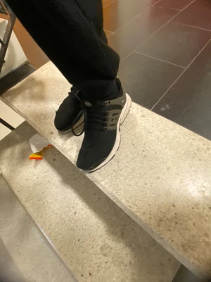 Nike Presto svarta sneakers - Nike Presto sneakers i svart med grå detaljer och vit sula. Skorna har en rund tå, snörning och en syntetisk ovandel med mesh för extra komfort. Snyggt Nike-logga på sidan och tå. Perfekt för dig som gillar sportig och clean stil.