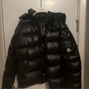 Svart Moncler pufferjacka med huva - Svart pufferjacka från Moncler med glansig finish, huva och ikonisk logga på ärmen. Jackan har dragkedja framtill, fickor med dragkedja och quiltad design. Insidan har en cool serietidningsetikett. Perfekt för kalla dagar och riktigt snygg streetstil.
